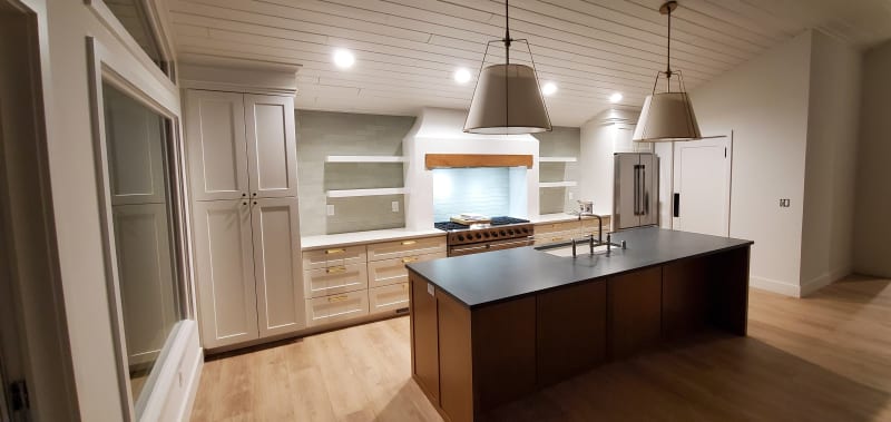 A beautifully remodeled kitchen by NIB in the Spokane, WA area. It features a spacious layout with modern finishes such as a sleek island with dark countertops, white cabinetry with gold hardware, and elegant light fixtures that add warmth to the space. The backsplash in soft green tones complements the overall design, creating a fresh and contemporary vibe. The kitchen is well-lit with recessed lighting and pendant lights over the island, creating a welcoming atmosphere perfect for cooking and entertaining.