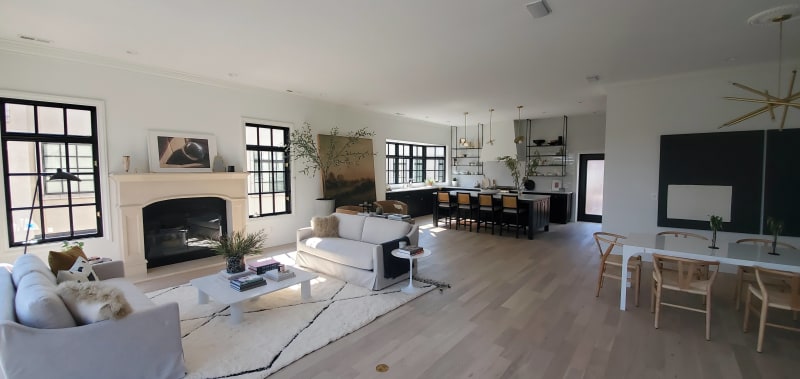 This NIB remodeling project in Spokane, WA, showcases a bright and modern living space with a seamless open floor plan. The living area features a cozy sofa arrangement centered around a sleek fireplace. The adjacent kitchen, with dark cabinetry, gold accents, and floating shelves, adds sophistication to the home. The space is bathed in natural light from large windows, with carefully curated decor adding to the stylish ambiance. The smooth wood flooring throughout complements the clean lines and light tones, creating an inviting atmosphere perfect for family gatherings and entertaining.
