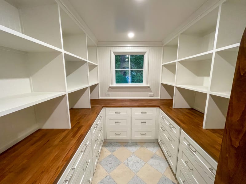 This NIB remodeling project in Spokane, WA, highlights a custom walk-in closet with an elegant design featuring white built-in shelving and drawers. The rich wood countertops add warmth to the space, creating a beautiful contrast with the white cabinetry. The shelving provides ample storage, while the well-lit environment, aided by natural light from the window, makes the space feel open and airy. The checkerboard marble flooring further enhances the sophisticated look of the closet, making it both functional and visually appealing.