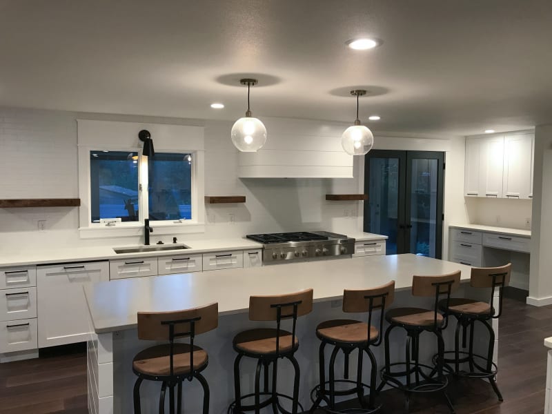 This kitchen remodel by NIB in Spokane, WA, features a bright and modern design, with an open layout that enhances both functionality and style. The expansive island provides ample space for meal prep and casual dining, complemented by the sleek white countertops and custom cabinetry. The high-end gas range, matte black faucet, and globe pendant lights contribute to the sophisticated yet relaxed atmosphere of the space. Large windows allow for abundant natural light, creating a welcoming environment perfect for both cooking and entertaining.