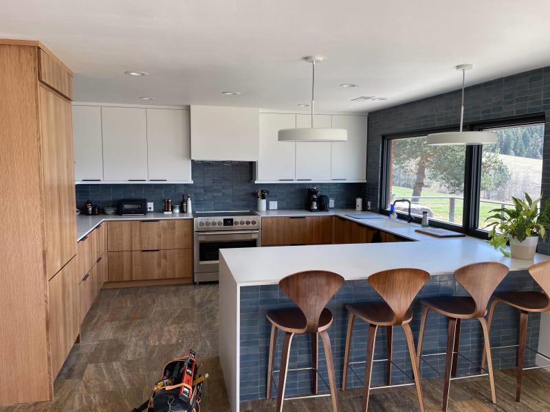 This beautifully built kitchen remodel in Spokane, WA, by NIB combines modern elegance with functionality. The striking contrast between the warm wood cabinetry and the deep blue tiles creates a sophisticated, yet inviting atmosphere. Large windows allow for a picturesque view of the surrounding landscape while natural light floods the space, highlighting the sleek countertops and minimalist design. The carefully selected light fixtures add a touch of charm to the open kitchen, making it the perfect space for cooking and entertaining.