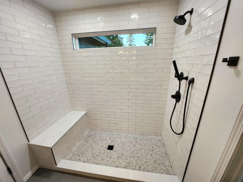 This stunning walk-in shower, completed by NIB in Spokane, WA, features clean, modern design elements. The sleek white subway tiles cover the walls, with a large window that allows natural light to brighten the space. A marble bench offers both style and comfort, while the hexagonal mosaic tile flooring adds a touch of elegance. The matte black fixtures provide a striking contrast to the light tiles, enhancing the overall aesthetic of the bathroom.