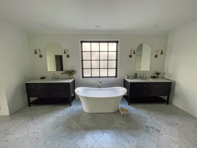 This beautiful bathroom remodel by NIB in Spokane, WA, showcases a modern, yet timeless design. The freestanding bathtub, placed centrally in the room, creates a luxurious focal point. Dark wood vanities, paired with marble tile flooring, offer a sophisticated contrast against the bright, airy walls. The large window provides natural light, enhancing the serene atmosphere of the space. This is a perfect example of how thoughtful design can blend functionality and style in a highly effective and visually stunning way.
