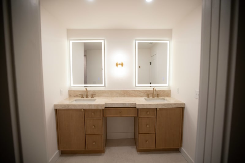 This modern bathroom remodel, completed by NIB in Spokane, WA, features a sleek, minimalist design. The elegant double vanity, with its clean lines and natural wood finish, creates a warm and inviting atmosphere. Framed by illuminated mirrors and paired with sophisticated gold faucets, the space exudes luxury and simplicity. This project emphasizes high-quality materials and subtle design elements, transforming the bathroom into a tranquil retreat.