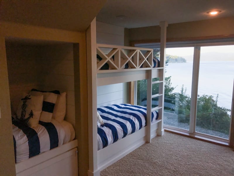 This cozy bunk room, created by NIB in Spokane, WA, offers a perfect space for relaxation and sleep with beautiful views of the water. The custom-built bunks are a standout feature, with crisp white frames and nautical-themed bedding that evoke a tranquil, seaside feel. Large windows let in plenty of natural light, highlighting the peaceful waterfront surroundings. Whether for family fun or restful nights, this room offers both charm and comfort in a serene setting.