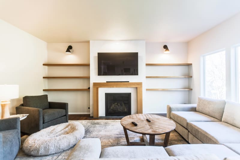 This elegant living room, remodeled by NIB in the Spokane, WA area, features a clean and modern aesthetic with a custom fireplace surround. The light-colored walls and plush sofas create a bright, airy space, while the natural wood shelving and earthy tones add warmth and texture. A carefully selected area rug complements the soft furnishings, and the strategically placed lighting accentuates the beautiful fireplace. The open shelving offers the perfect opportunity to display personal décor, completing the inviting atmosphere of this thoughtful renovation.