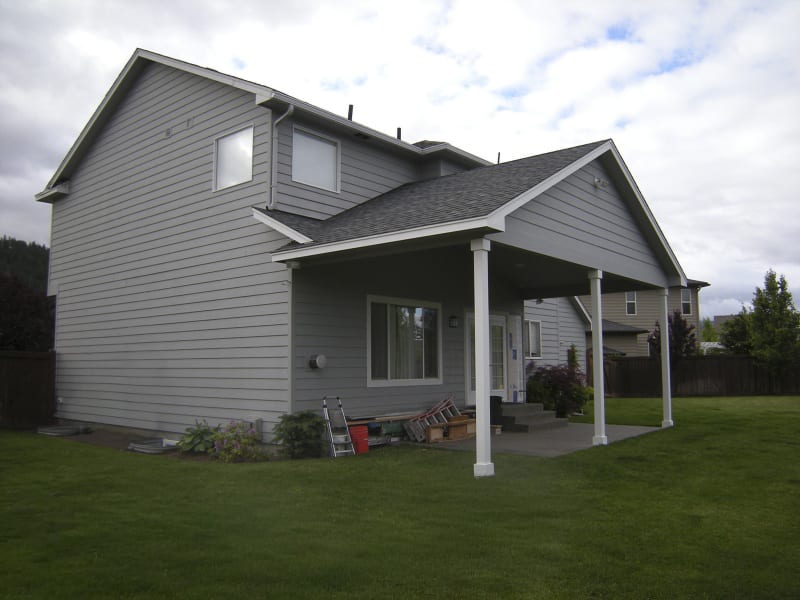 A residential property with a functional outdoor space. The well-maintained backyard features a patio with a covered area supported by two sturdy white columns, providing a sheltered spot for relaxation. The gray siding of the house complements the lush green lawn and tidy landscaping, creating a comfortable and inviting environment. The surrounding privacy fence enhances the peaceful atmosphere, making this outdoor area a perfect retreat in Spokane, WA.