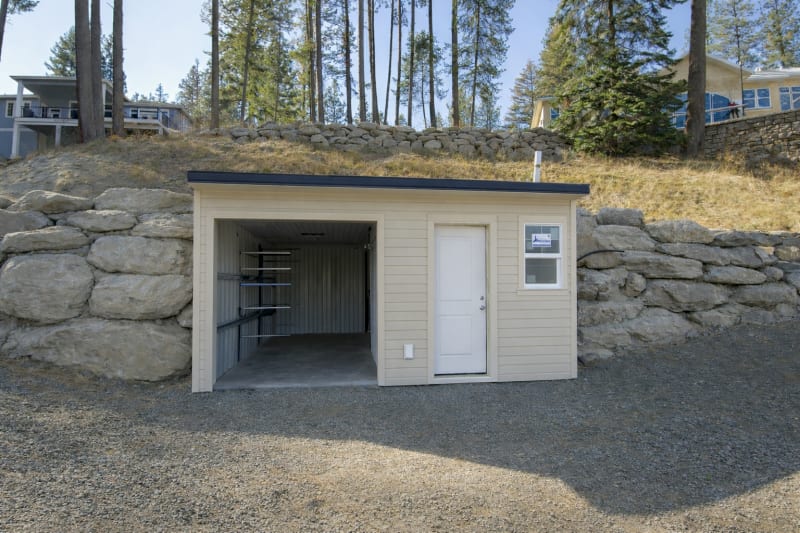 A custom-built storage shed designed and constructed by NIB in Spokane, WA. The shed, nestled against a natural rock formation, offers ample space for tools, equipment, and other outdoor essentials. The simple, yet sturdy design features a clean exterior with a single door and window. The gravel base and surrounding landscape complement the structure's minimalist style, making it a practical and reliable addition to any outdoor space.