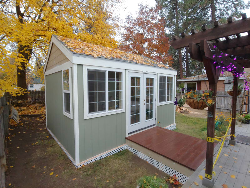 A beautifully crafted outdoor space in Spokane, WA. The focal point is a small, well-constructed shed with large windows and a wooden deck at its entrance, providing a charming touch to the backyard. The surrounding fall colors add vibrancy to the scene, with golden leaves scattered across the roof. A decorative pergola with purple lights and a well-maintained path leading to the shed further enhance the tranquil ambiance of the outdoor area, making it a perfect functional retreat.