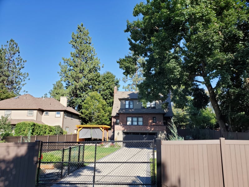 A charming home in Spokane, WA, featuring a functional outdoor space. The property is enclosed by a combination of a sturdy brown privacy fence and a black chain-link gate that adds both security and curb appeal. In the backyard, an elegantly designed pergola stands as a focal point, offering a perfect space for outdoor relaxation or gatherings. The well-maintained lawn and surrounding trees create a tranquil and inviting environment.