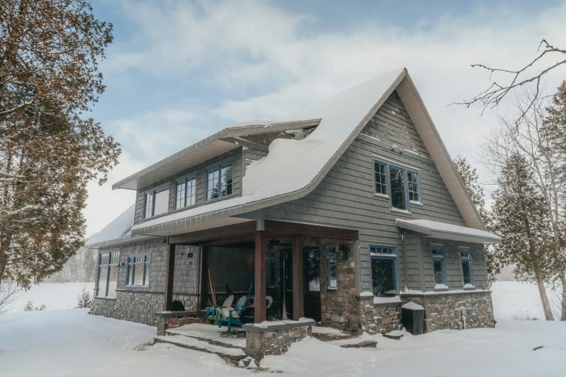 Beautiful shot of a home. The home's exterior features a combination of shingles and stonework that blends seamlessly with the snowy landscape, giving it a warm, rustic charm despite the winter chill. The careful attention to detail in the siding installation and choice of materials enhances the home's overall aesthetic while providing a durable finish built to withstand the elements. 