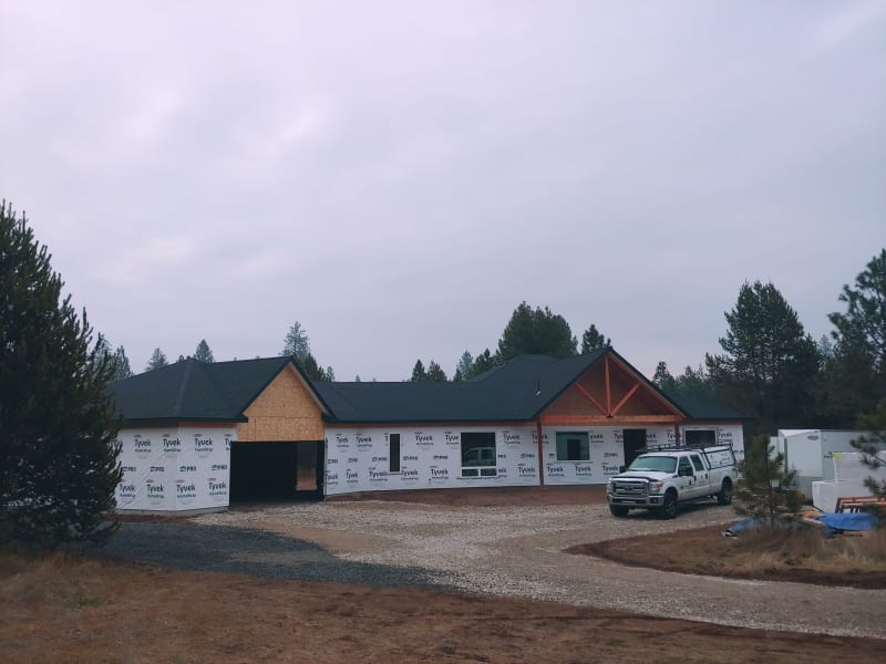 A custom home being framed with Tyvek house wrap installed, part of an NIB custom home build project in the Spokane, WA area. The roof is in place, but the garage remains open for further work. A construction vehicle is stationed nearby, and the setting is surrounded by trees.