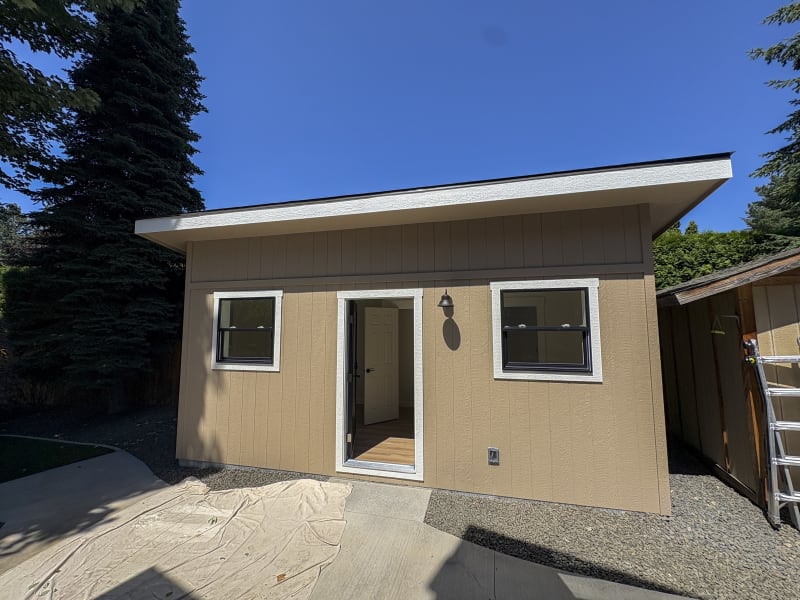 A newly constructed structure featuring fresh siding/roofing/soffit/fascia installation by NIB in the Spokane, WA area. The exterior features clean lines with tan-colored siding and a well-maintained gravel base around the structure. Windows are neatly framed, and the door is centered, contributing to the simple yet modern aesthetic. The surrounding environment includes greenery, and the overall look is fresh and inviting.