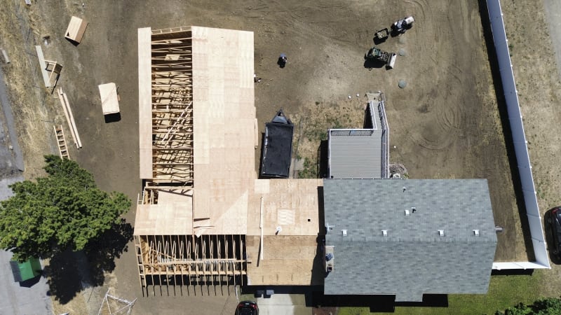 Wooden framework of an attached custom home addition under construction, captured from an overhead perspective. The beginning stages, with the main structure extending out from the existing building. The groundwork is visible, with materials like plywood and equipment scattered across the lot. This project is being built by NIB in Spokane, WA.