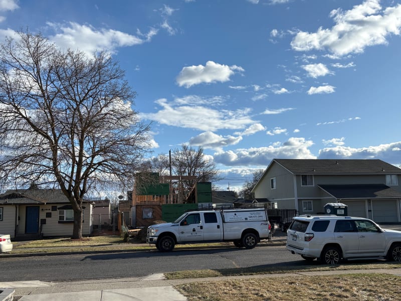 A construction site for a custom home is visible in the foreground, with framing work underway. NIB's truck is parked out front, and nearby homes are visible in the background. The scene shows a bright day with scattered clouds over Spokane, WA, where NIB is carrying out the construction work.