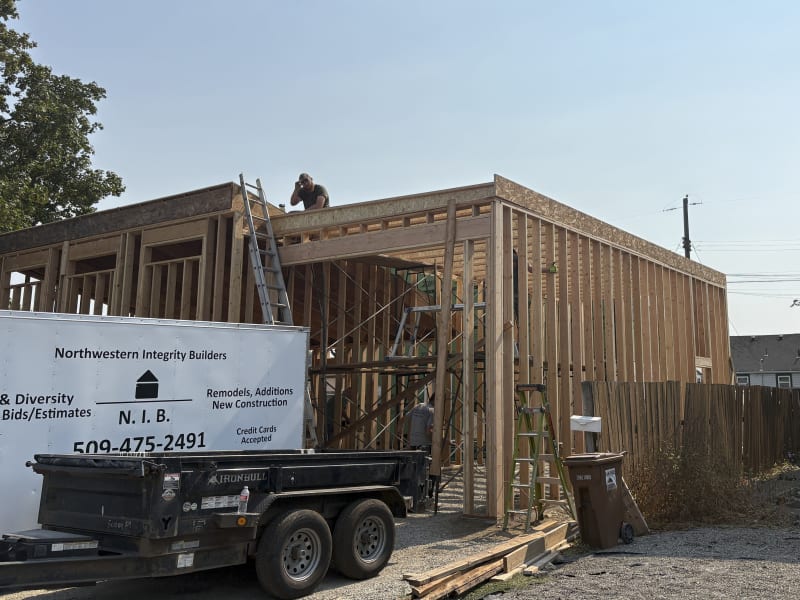 A partially framed home addition is in progress, with a worker on the roof. The image includes a ladder, scaffolding, and equipment, such as a trailer and a garbage bin. The project, completed by NIB in Spokane, WA, demonstrates the early stages of building a home extension.