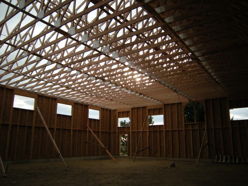 Interior of a large horse barn under construction, showcasing the wooden framework of the ceiling and walls. The roof trusses are visibly in place, creating an open, airy feel, with natural light filtering through the gaps. The project is in its framing phase, and the barn's structure is taking shape, with supports and braces strategically placed to stabilize the frame as the next steps of construction proceed. This setting demonstrates the precision and skill involved in building large, functional spaces with an eye for structural integrity.