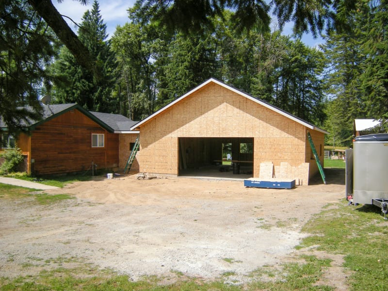 Custom home construction project in Spokane, WA, in the early stages of framing. The garage portion of the building has been framed and the exterior wall is covered with OSB sheathing. The area around the home is still being leveled, and the worksite includes construction tools such as ladders and a trailer. The surrounding environment consists of lush trees and a gravel driveway, highlighting the property's scenic location. NIB's craftsmanship is evident as the home begins to take shape.