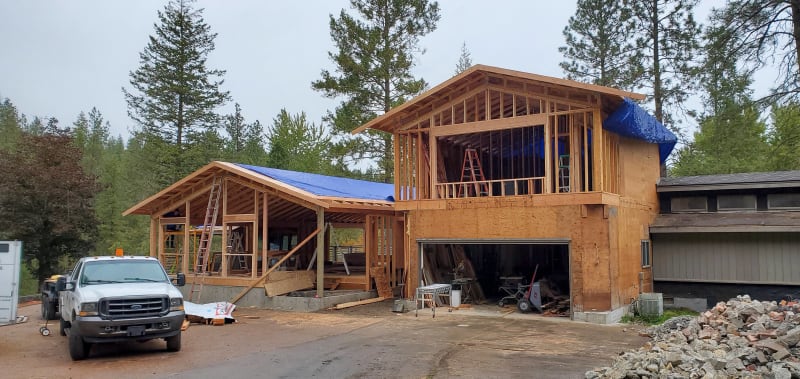 A custom home being built in Spokane, WA, by NIB with significant progress on its framing. A large section of the roof structure has been completed, while some areas remain covered with protective blue tarps to safeguard the materials. The worksite is equipped with ladders, tools, and construction vehicles, with a clear view of the surrounding landscape. NIB’s team continues to manage the project, ensuring each stage is carefully executed, from foundational elements to roofing, providing quality craftsmanship for the future homeowners.