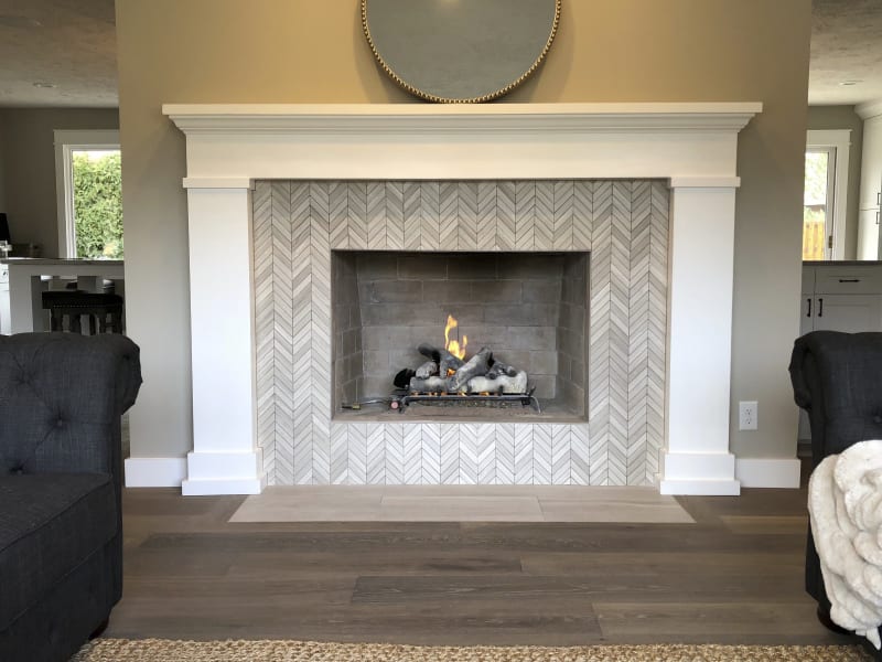 A beautifully designed living room fireplace in the Spokane Valley area of Spokane, WA. The fireplace features a striking herringbone-patterned tile surround in soft neutral tones, creating a sophisticated focal point. The white trim and mantle frame the fireplace, complementing the clean lines of the room. A modern gas fire glows gently inside, adding warmth and ambiance to the space. The surrounding hardwood flooring and cozy furniture complete the inviting atmosphere, making this a perfect living room centerpiece.