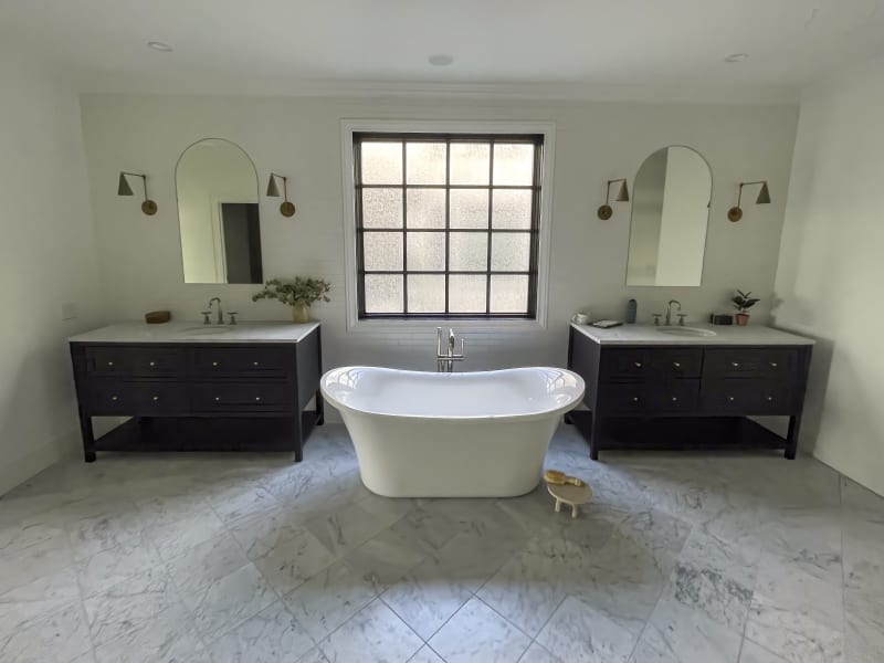 A stylish bathroom remodel in the Spokane Valley area of Spokane, WA. The space is centered around a sleek freestanding bathtub, positioned between two dark wood vanities. Each vanity is topped with white marble countertops and complemented by round, brass-framed mirrors and modern wall sconces. A large window allows natural light to illuminate the clean, bright space, with subtle decorative plants adding a touch of greenery. The marble flooring extends throughout the room, contributing to the sophisticated and timeless design.
