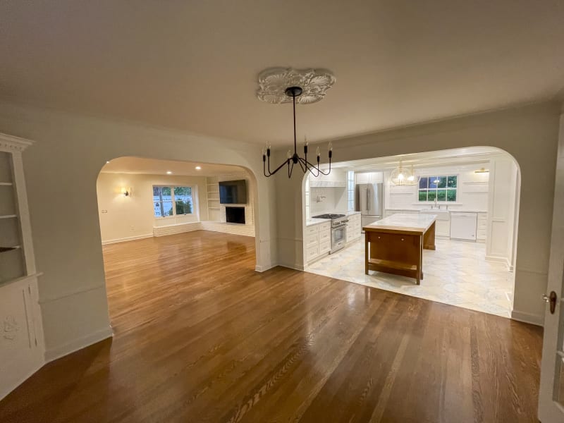 A beautifully remodeled living and kitchen space in the Spokane Valley area of Spokane, WA. The living room, visible through an arched doorway, features hardwood flooring, a built-in bookshelf, and a cozy fireplace. The kitchen on the other side is spacious and modern, with white cabinetry and a large central island. The countertops are made of light-colored marble, complementing the stainless steel appliances and bright lighting. The open-concept design highlights the seamless flow between the two areas, with large windows letting in natural light.
