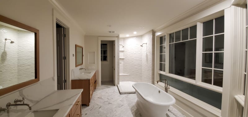 A beautifully remodeled bathroom in the Spokane Valley area of Spokane, WA. The spacious room features a freestanding white bathtub positioned next to a large window, allowing natural light to flood the space. The bathroom is finished with sleek white marble flooring in a herringbone pattern, and the walls are covered in light-colored stone tiles. A modern vanity with a marble countertop and wood cabinetry complements the clean, minimalist aesthetic of the space. The shower area is equipped with elegant fixtures and built-in shelving.