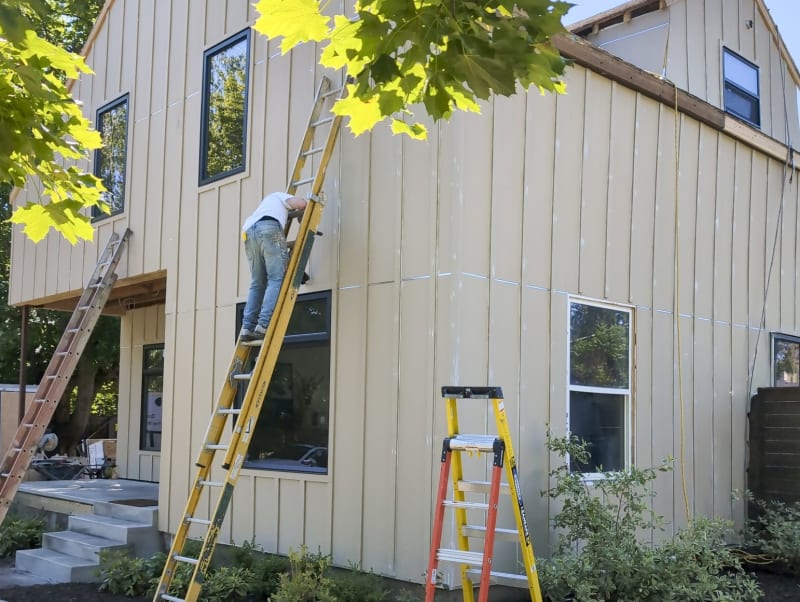 A siding installation is in progress in the Moran Prairie area of Spokane, WA. Two ladders are positioned against the exterior of a newly constructed home, where NIB workers are applying siding panels to the upper floors. The light beige siding contrasts nicely with the large windows, and the clean lines of the installation are visible. The lush greenery from surrounding trees adds to the natural aesthetic of the worksite, which also shows steps leading up to the front door.