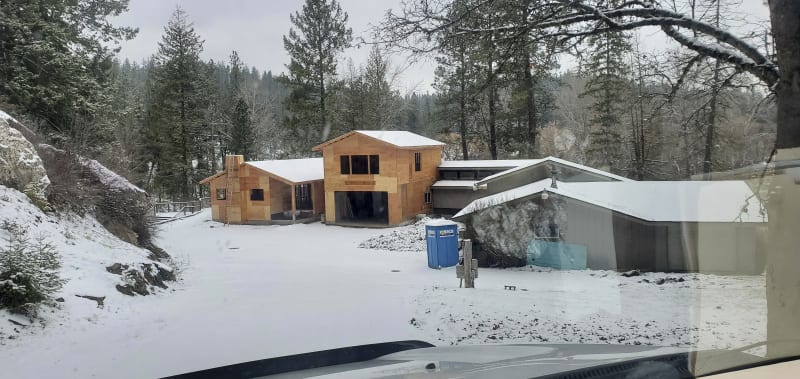 A new home additions construction project in the Moran Prairie area of Spokane, WA, during winter. Exteriors are covered with plywood sheathing, and the structures are taking shape with framing visible. Snow blankets the ground and rooftops, adding a serene winter backdrop to the site. A ladder rests against the side of the building, and a portable toilet is placed nearby. The surrounding forested landscape enhances the secluded, rural atmosphere of the worksite.