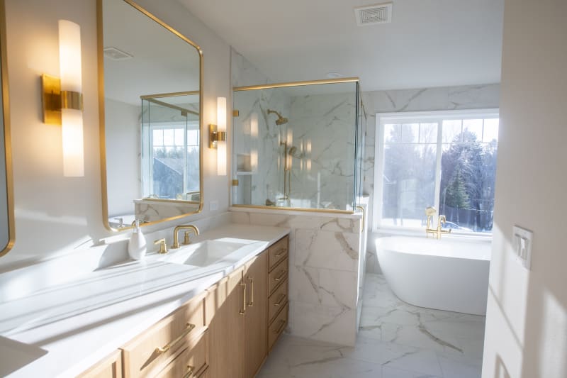 A luxurious bathroom remodel in the Moran Prairie area of Spokane, WA. The space features a modern vanity with light wood cabinetry and elegant gold fixtures, paired with a white marble countertop. A spacious walk-in shower, encased in clear glass, boasts marble tiles with golden accents. A freestanding tub sits beneath a large window, offering natural light that floods the room. Soft lighting from sconces mounted beside the mirrors enhances the sophisticated, serene atmosphere of the bathroom.