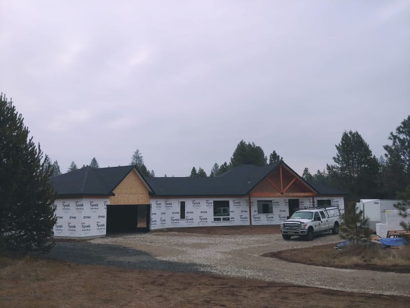 A home construction project in the Moran Prairie area of Spokane, WA, in its early stages. The exterior is wrapped in Tyvek house wrap, and the roof is fully installed with dark shingles. The front of the home features a rustic wooden gable detail, adding character to the design. A white NIB work truck is parked in the driveway, and construction materials are scattered around the site. The surrounding landscape is sparse with dry grasses, trees, and a cloudy sky, giving the scene a quiet, overcast ambiance.