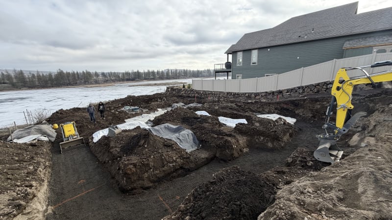 A home construction site in the Liberty Lake area of Spokane, WA, showing the early stages of excavation for a new project. The site features a series of trenches and mounds of dirt, some covered with tarps to protect the ground. A small construction vehicle is actively working, moving dirt along a cleared path. In the background, a residential home with a fenced yard sits beside a frozen lake, creating a tranquil and scenic backdrop for the ongoing construction work.