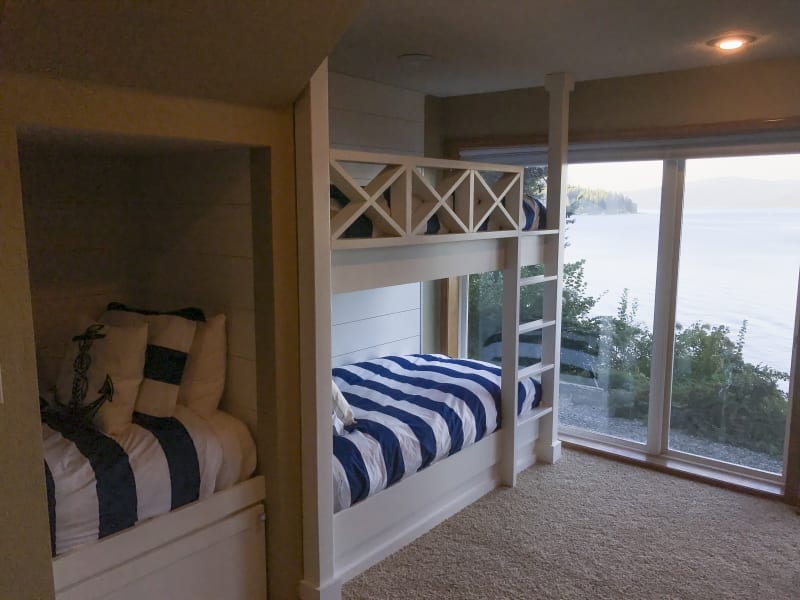 A charming bunk room in the Liberty Lake area of Spokane, WA, featuring a nautical-inspired design. The bunk beds are crafted with white wood, with one bed placed underneath the other and separated by a sleek ladder. Each bed is outfitted with striped navy and white bedding, complementing the room’s coastal theme. The large window behind the beds offers a stunning view of the lake, bringing natural light and scenic beauty into the space. The soft carpet and clean, simple design create a cozy and inviting atmosphere.