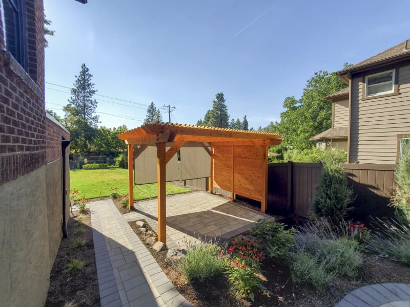 A beautifully designed pergola in the Liberty Lake area of Spokane, WA, in a serene backyard. The structure features natural wood with a slatted roof that offers partial shade, creating a comfortable outdoor living space. The pergola is surrounded by lush greenery, including colorful flowers and shrubs along a stone pathway that leads to it. The clean, modern fence and neatly manicured lawn add to the peaceful and inviting atmosphere of this outdoor retreat.