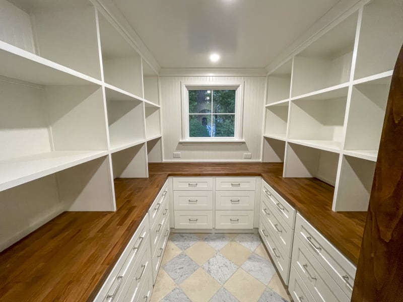 A beautifully designed walk-in closet remodel in the Fairwood area of Spokane, WA. The space features ample white shelving, providing plenty of storage for clothes and accessories. Below the shelves, wooden countertops create a warm contrast, and sleek white drawers offer additional organization. The floor is finished with elegant marble tiles, enhancing the room's sophisticated feel. A large window allows natural light to brighten the space, creating a pleasant and functional environment for any homeowner.
