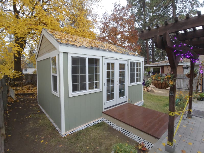 A charming backyard shed remodel in the Fairwood area of Spokane, WA. The structure is finished in soft green siding with white trim and features large windows, allowing plenty of natural light to enter the space. A small wooden deck with a set of French doors welcomes entry, while the surrounding yard is dotted with autumn leaves. A pergola with purple lights adds a decorative touch to the nearby area, complementing the cozy and functional vibe of the shed.