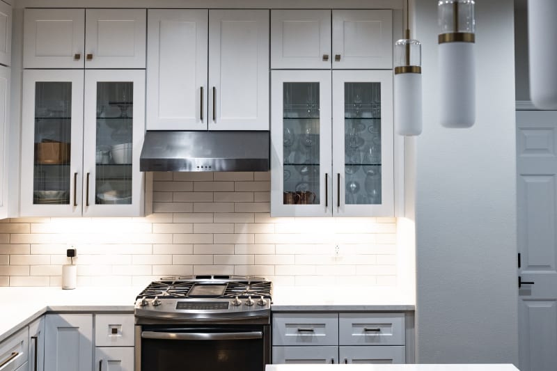 A cozy and stylish bedroom remodel in the Fairwood area of Spokane, WA. The room features a light neutral color palette with a feature wall of paneling behind the bed, adding depth and texture. The bed is dressed with soft bedding and decorative pillows in muted tones of pink and burgundy, giving the room a calm, welcoming feel. On either side of the bed are traditional nightstands with table lamps, creating a balanced, serene environment. The hardwood flooring and soft area rug complete the comfortable and timeless design.