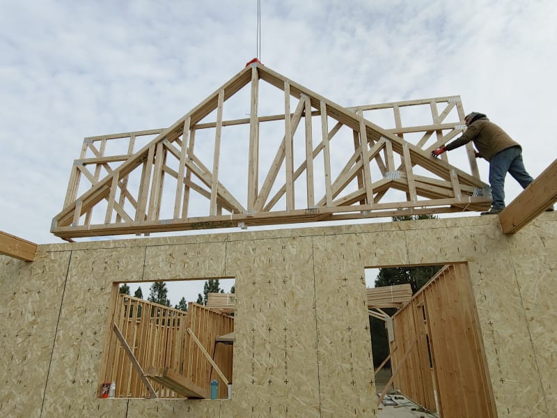 A home framing project in the Fairwood area of Spokane, WA, showing the installation of the roof trusses. One worker is positioning a large truss with the help of a crane, while another worker ensures everything is aligned properly. The building’s walls are framed with plywood sheathing, and a window opening is visible on one side. The sky above is partly cloudy, and the surrounding construction site is tidy, with tools and materials nearby.