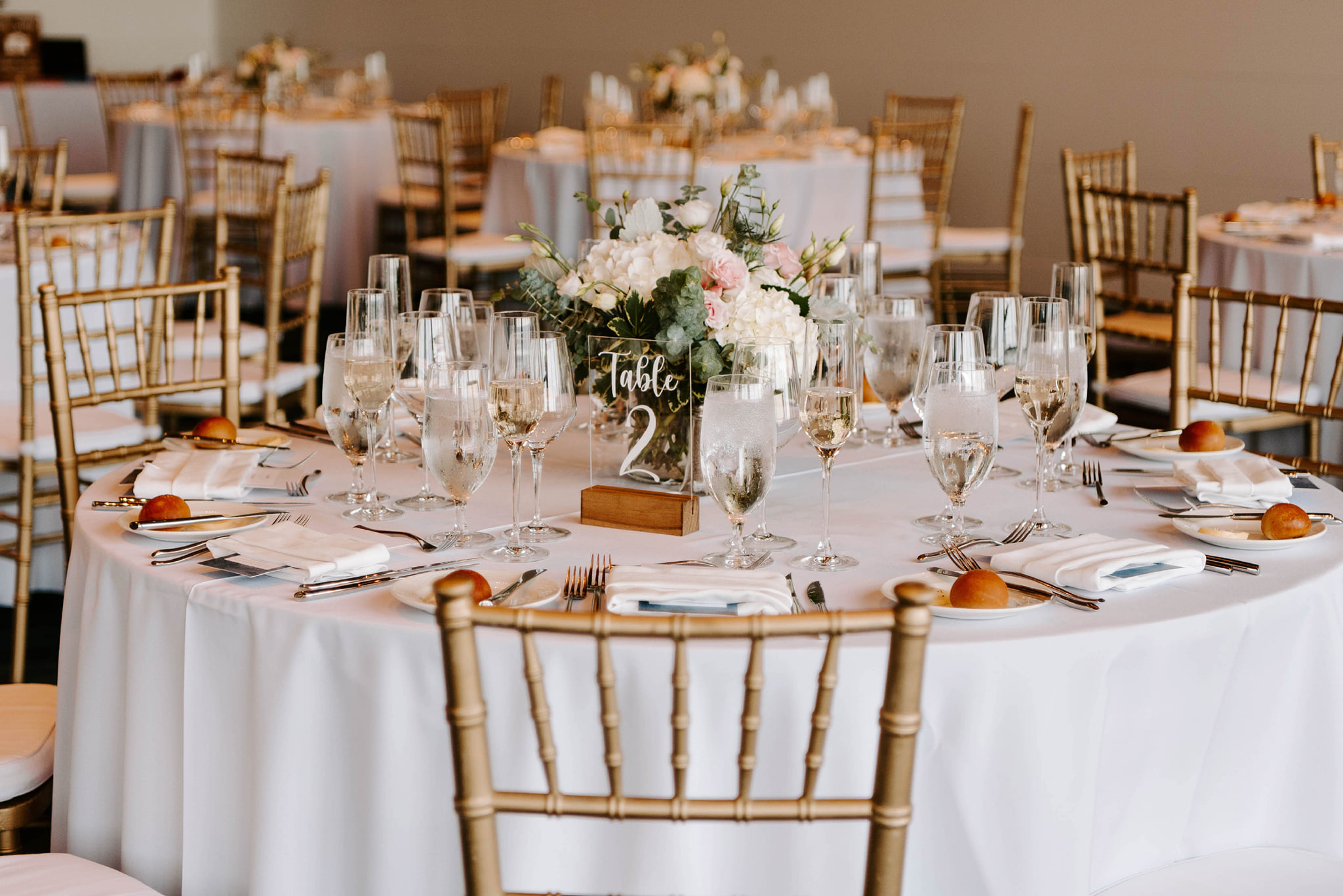 Wedding reception table with a white and blush floral centerpiece and gold chairs.