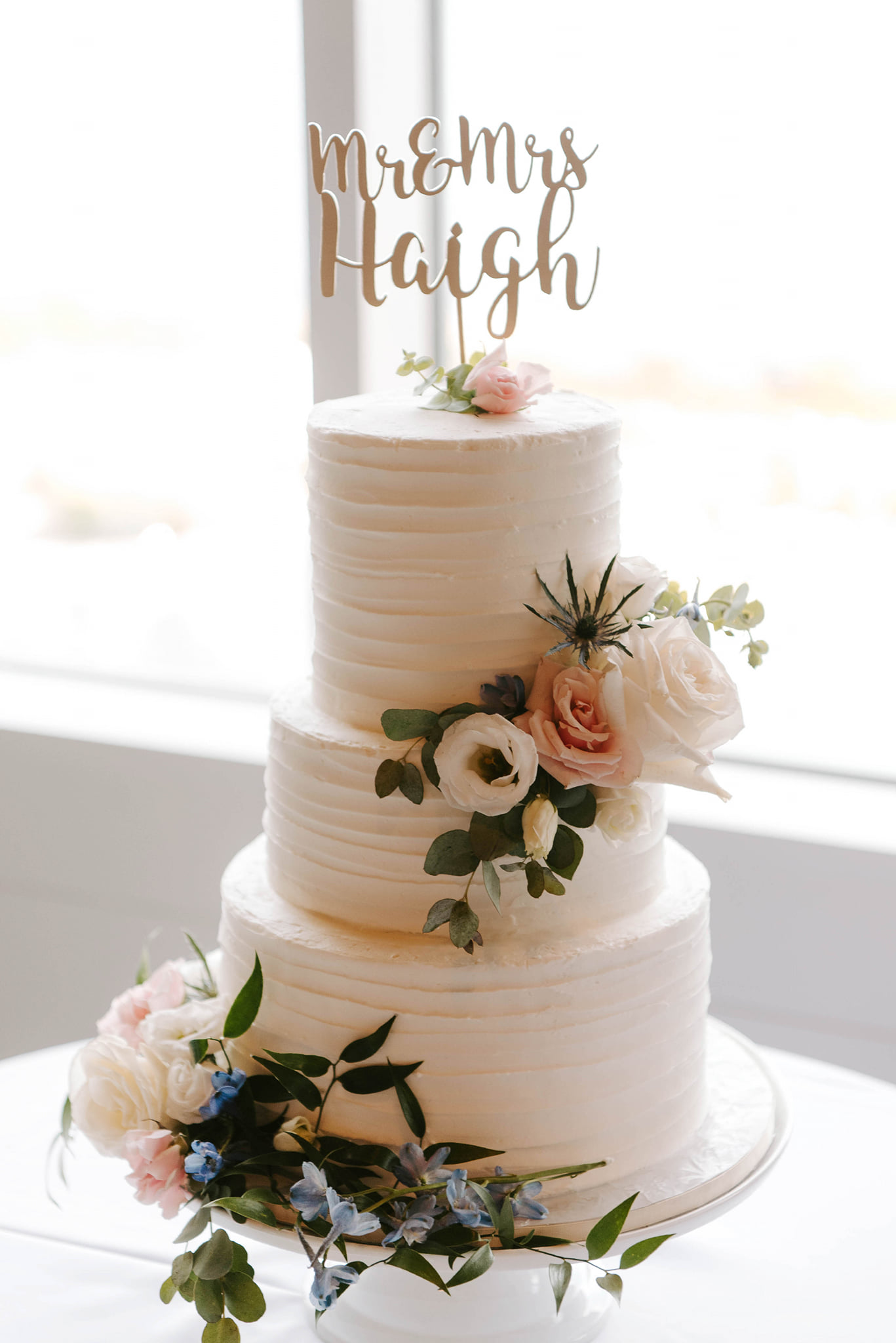 Wedding cake decorated with blush, white, blue flowers and fresh greenery.