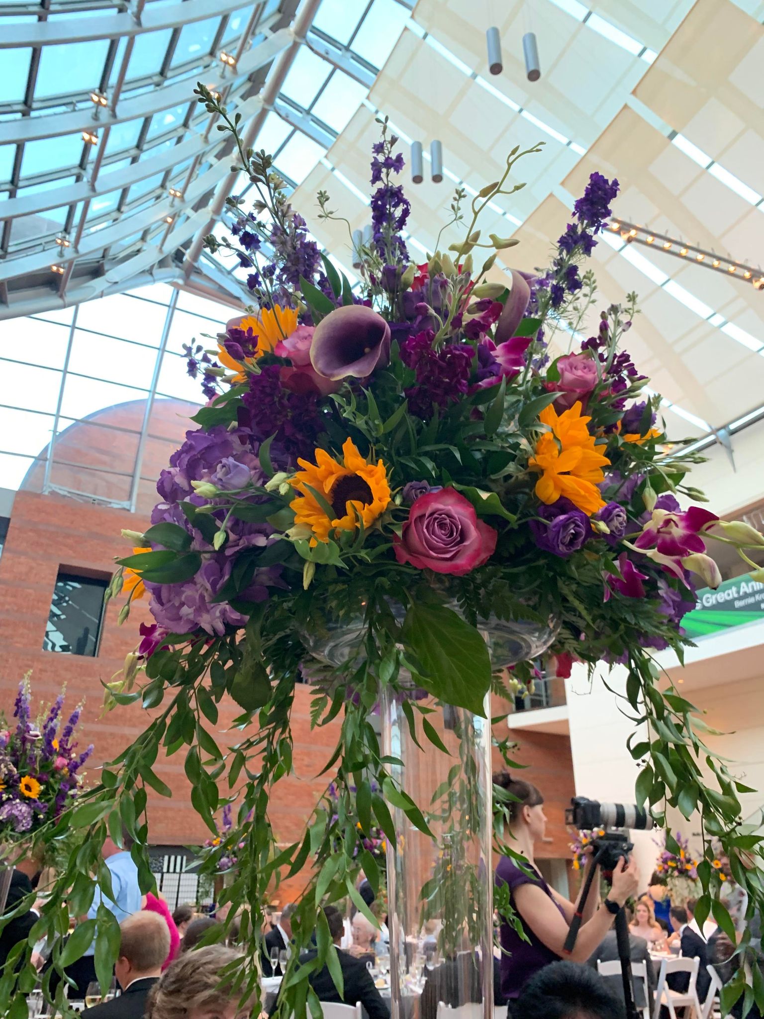 Tall wedding centerpiece with sunflowers, purple flowers, pink roses, orchids, and trailing greenery.