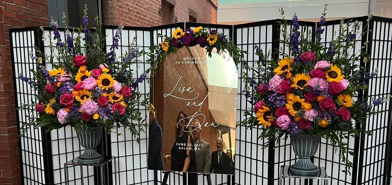 Wedding welcome sign framed by colorful floral arrangements with sunflowers, roses, purple flowers, and greenery.