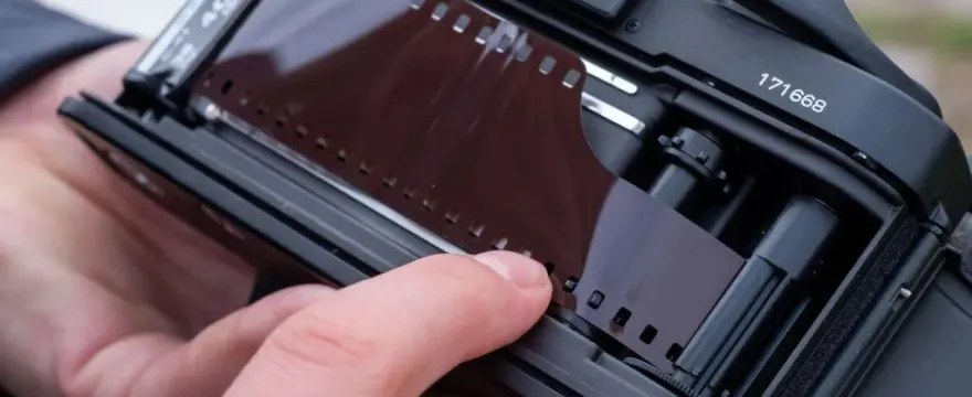 Hands loading a roll of 35mm film into an SLR camera, showing the film back open