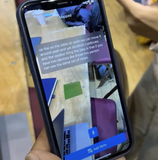 An iPhone being held with the camera open pointing at a table. On the table is a green post-it note. Overlaid is a text box with the current speech being displayed, and an 'add note' button at the bottom of the screen.