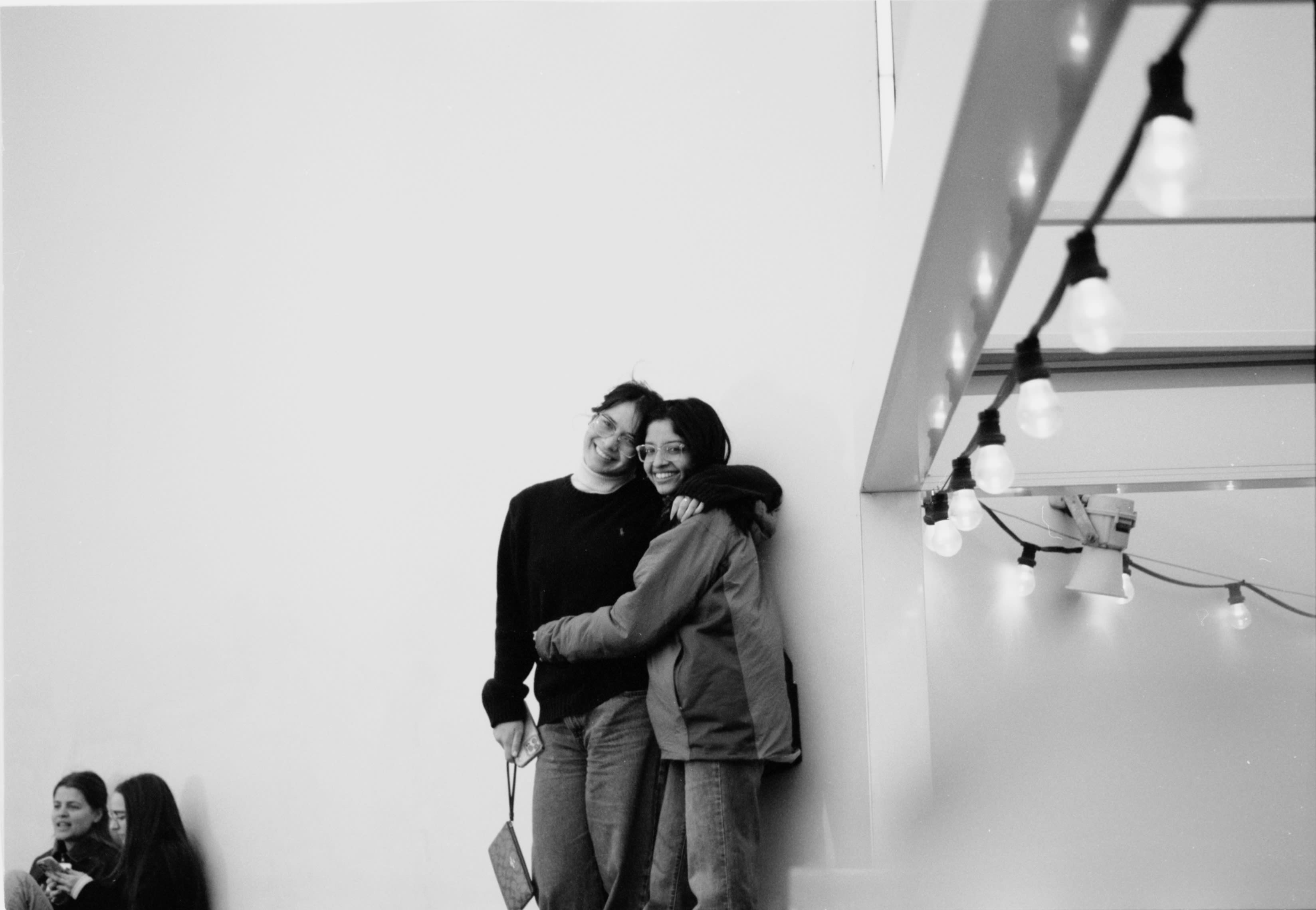 Two friends hugging against a white wall under string lights