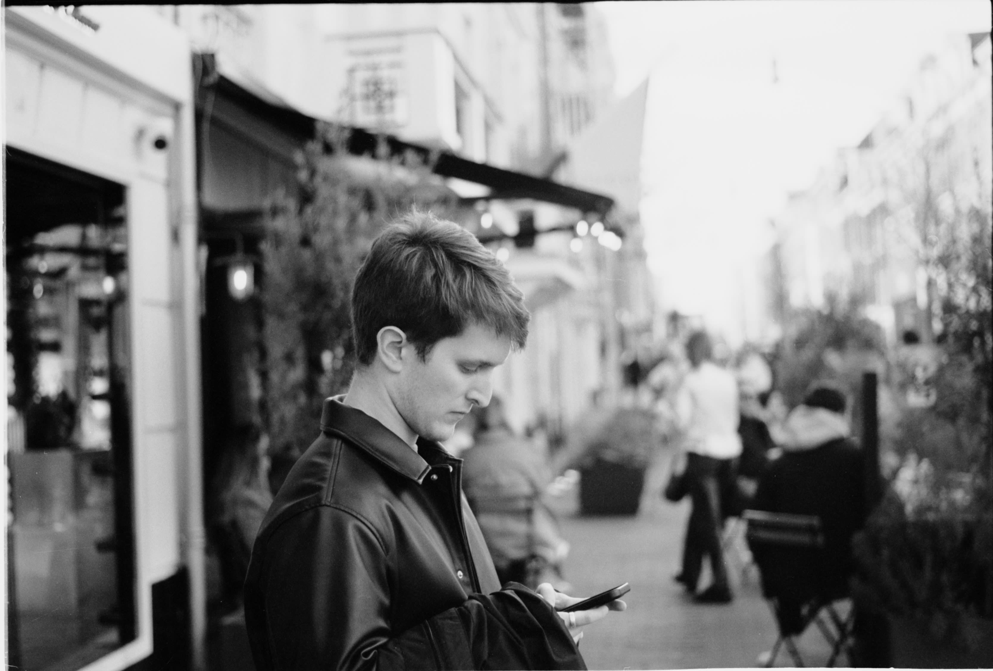 Someone in a leather jacket checking their phone on a street of cafés