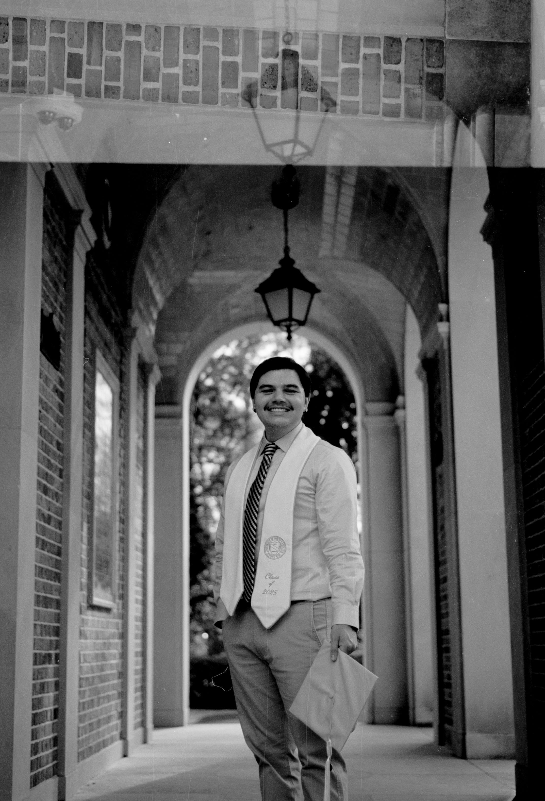 A grad holding a cap in an arched brick walkway under a hanging lantern