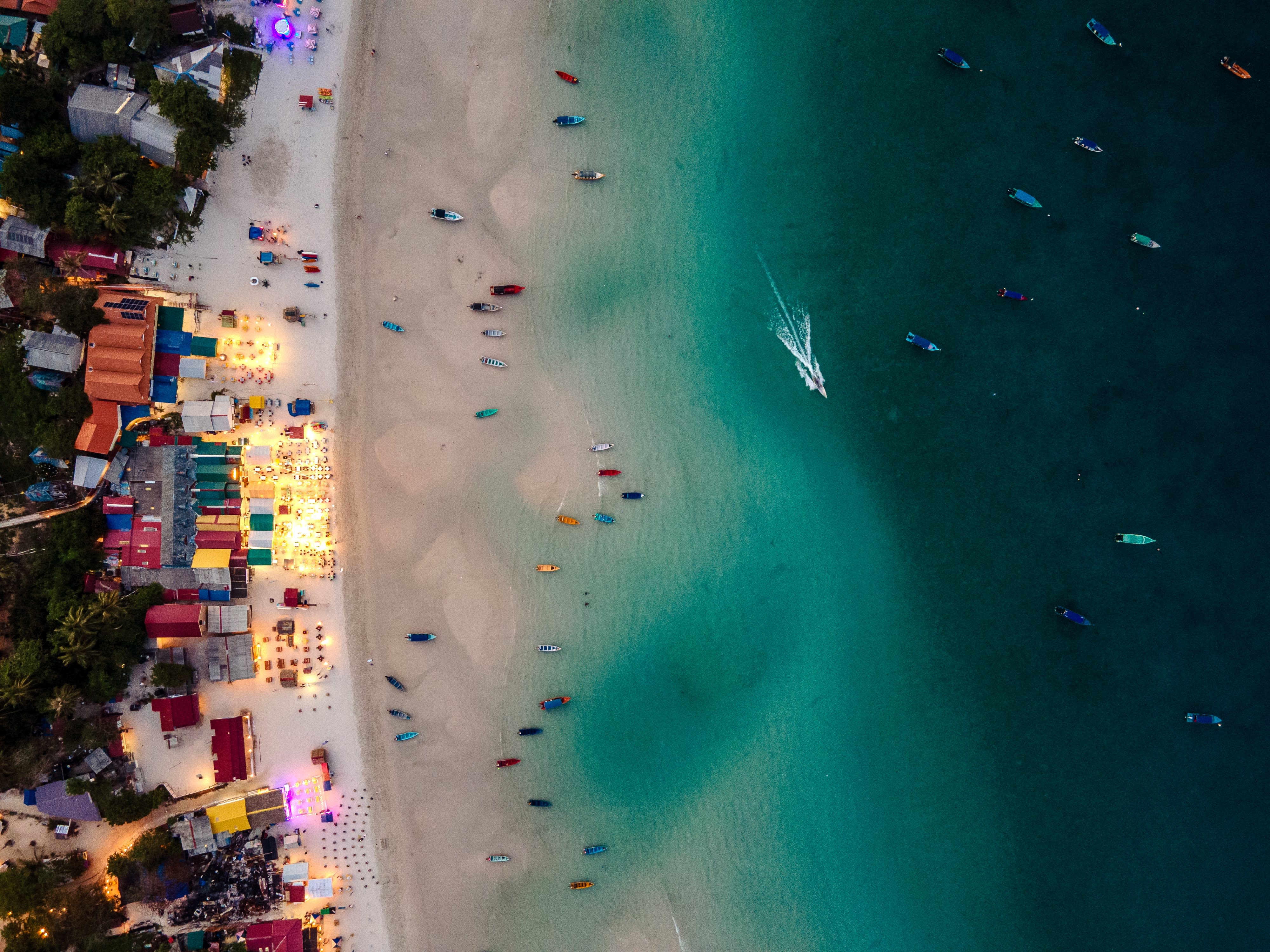 Long Beach at dusk from above: lit bars and colorful huts along the sand, boats moored in turquoise water