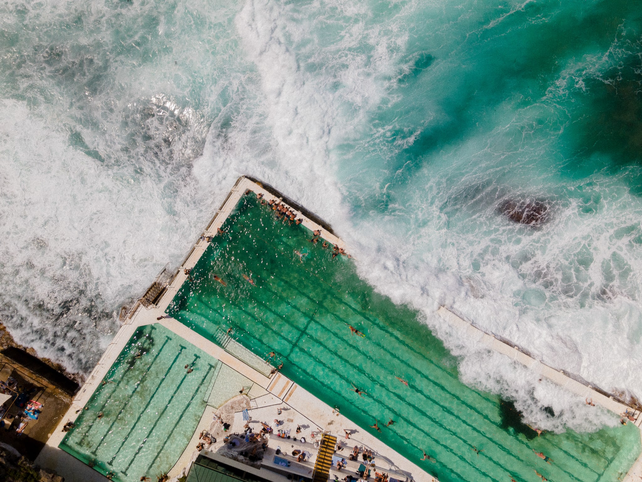Looking straight down at the Bondi Icebergs pool as waves break over its seaward wall, swimmers in the lanes
