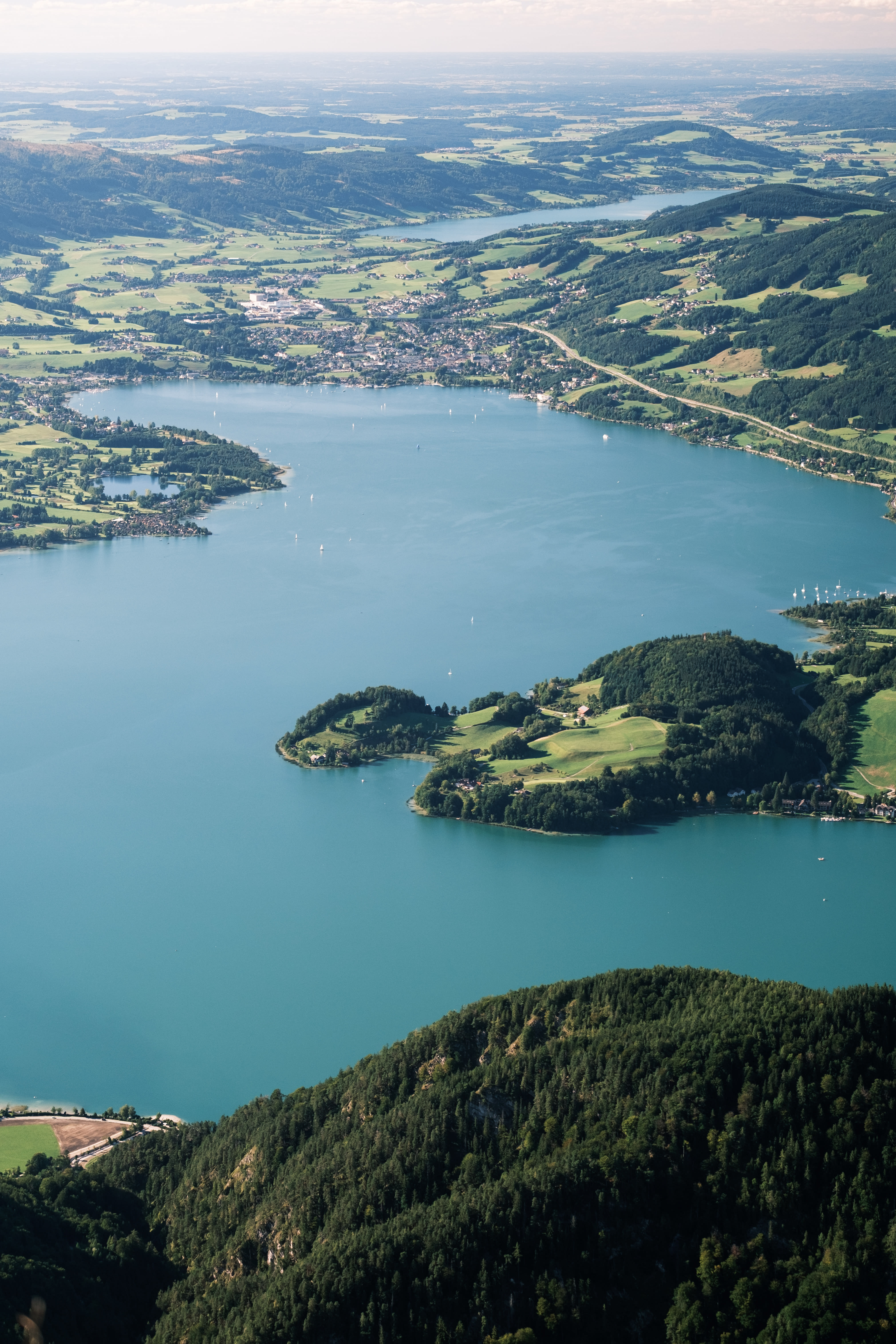 A long blue lake winding between green hills and villages, seen from a forested mountain